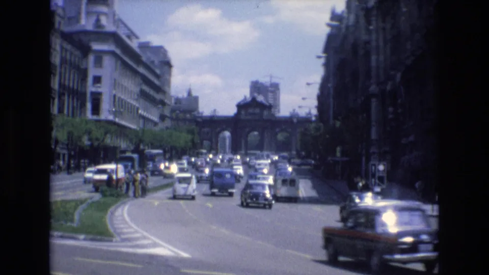 1972 Spain urban traffic scene with several classic cars, pedestrians near the curb and median, tall street-facing buildings, traffic signals, and a distant arched gateway.