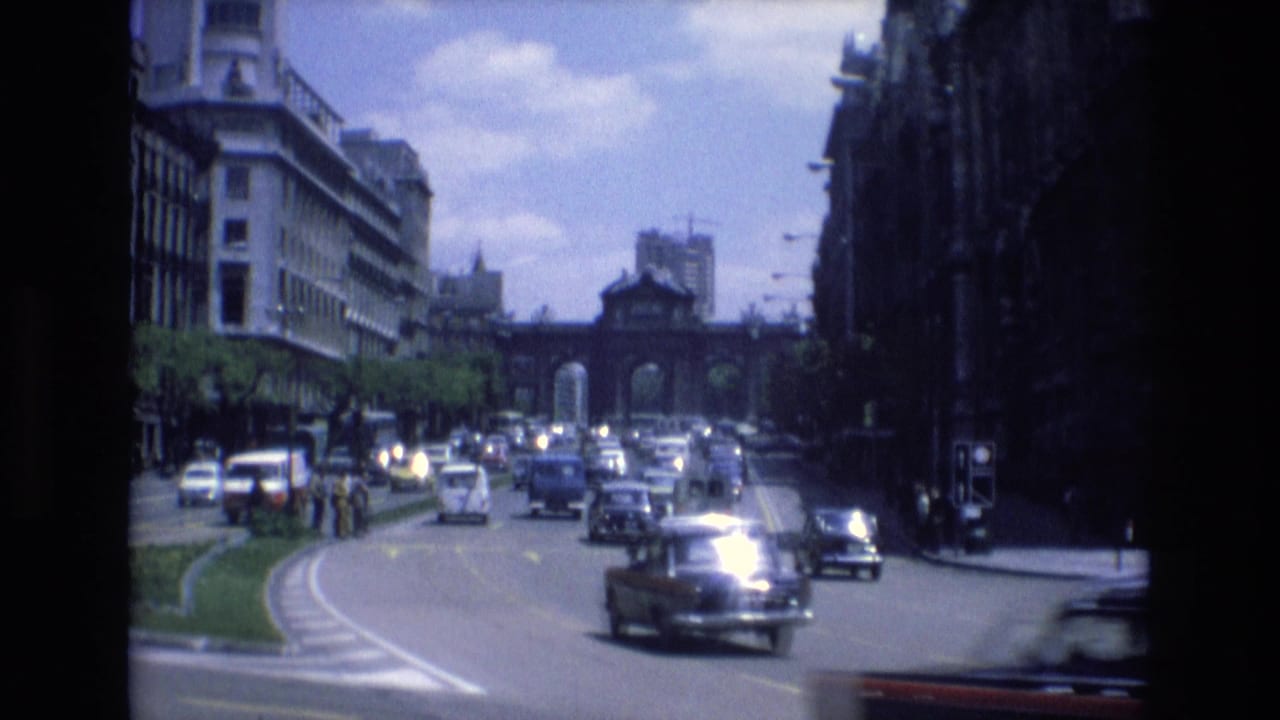 1972 Spain 8mm frame showing cars moving through a broad urban intersection with a planted median, sidewalks, and an arched gateway in the distance.