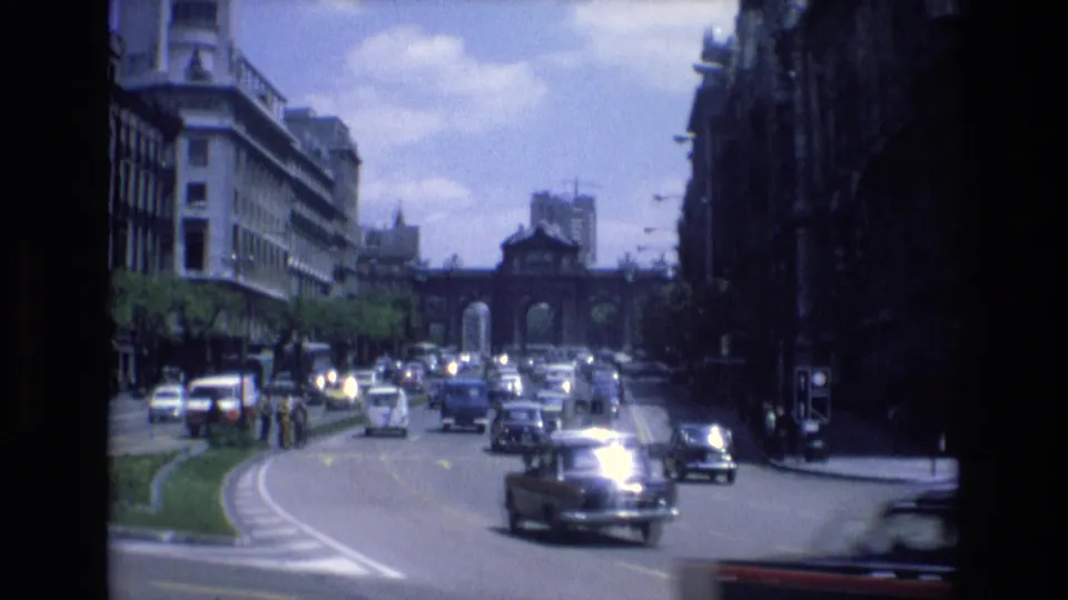 1972 Spain 8mm frame showing cars moving through a broad urban intersection with a planted median, sidewalks, and an arched gateway in the distance.
