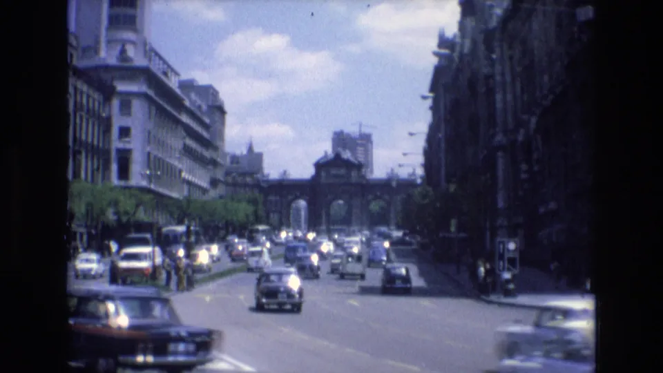 1972 Spain 8mm frame of cars passing through a sunlit city avenue, with dark building facades on one side and an arched gateway at the far end.