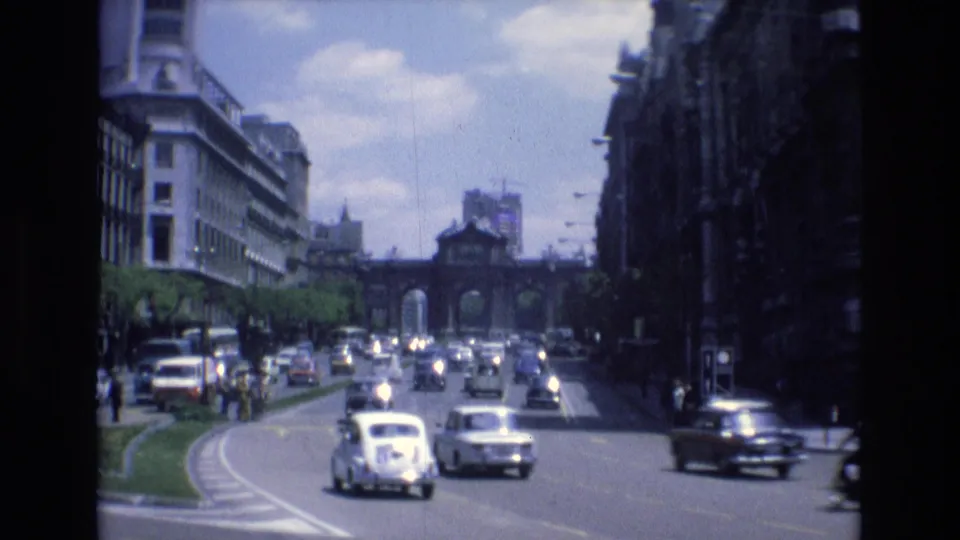 1972 Spain 8mm frame showing compact cars and sedans navigating a city roadway with a green median and distant arched gateway.