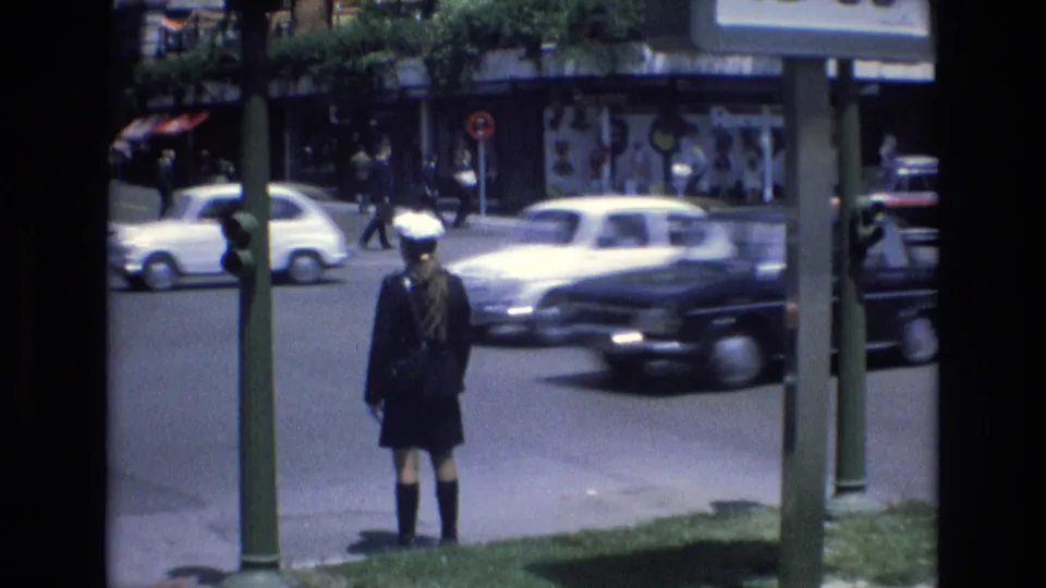 1972 Spain 8mm frame with a uniformed pedestrian standing near signal poles while cars pass across a busy urban intersection and storefronts line the background.