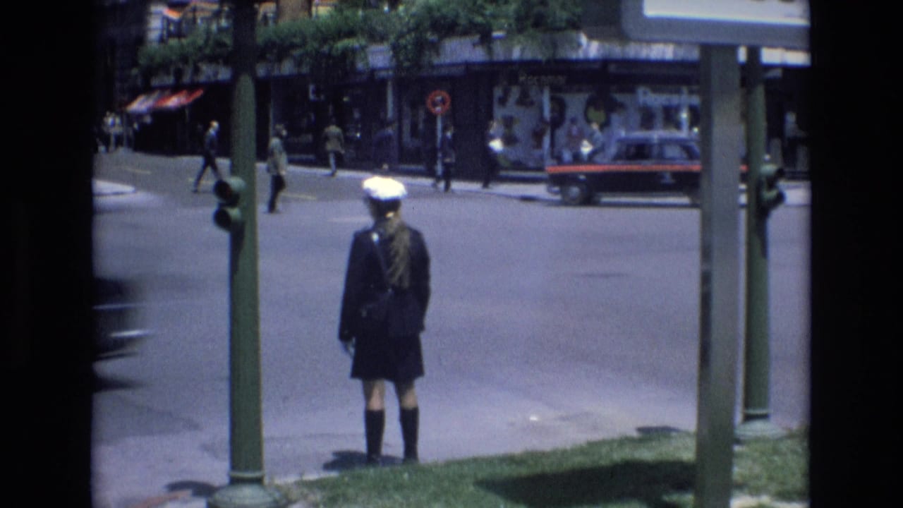 In 1972 Spain, a uniformed young figure seen from behind stands by green street poles at a sunny city intersection with pedestrians, storefronts, and traffic beyond.