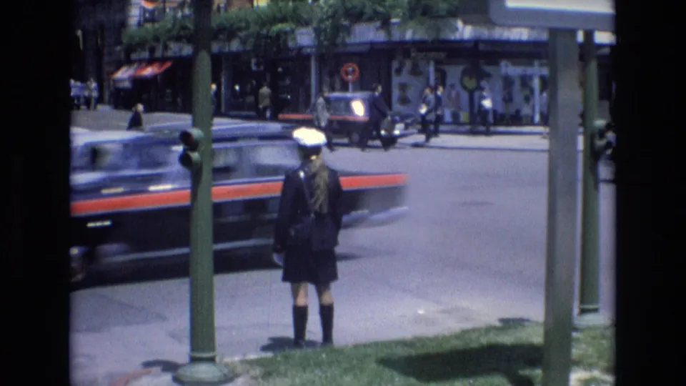 A dark car with a red side stripe blurs past the stationary uniformed figure at a Spanish city street corner, with pedestrians and shop windows visible across the road.