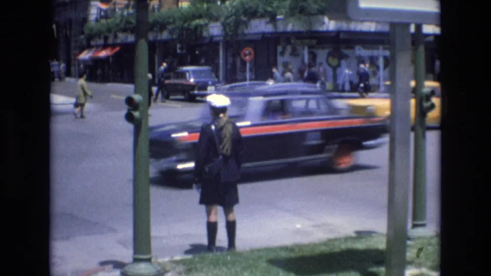 A blurred dark vehicle moves through the foreground as the uniformed figure stands by the curb, with storefronts, pedestrians, and another car visible across the intersection.