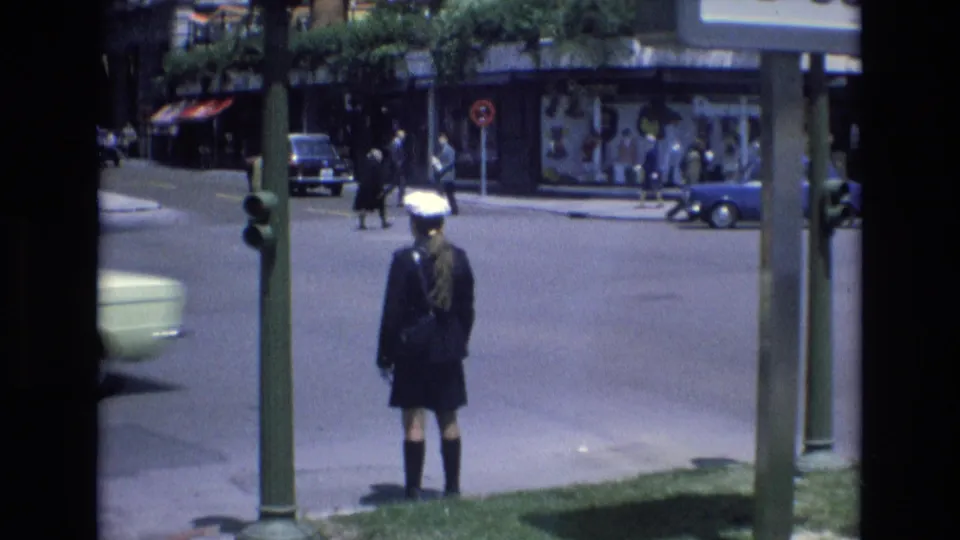 The final frame holds on the uniformed figure at the curb while a small light-colored car, a blue car, pedestrians, street poles, and storefronts remain visible in the 1972 Spain street scene.