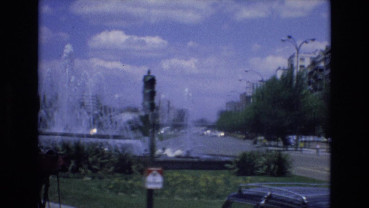 A 1972 Spain 8mm frame shows a large city fountain across a landscaped strip, with a road, cars, street lamps, and apartment buildings in the background.