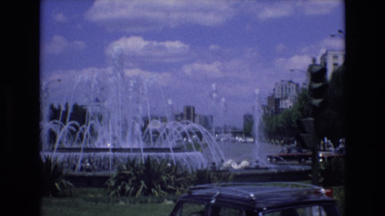 Active fountain with arcing water jets, greenery, traffic, and buildings behind it.