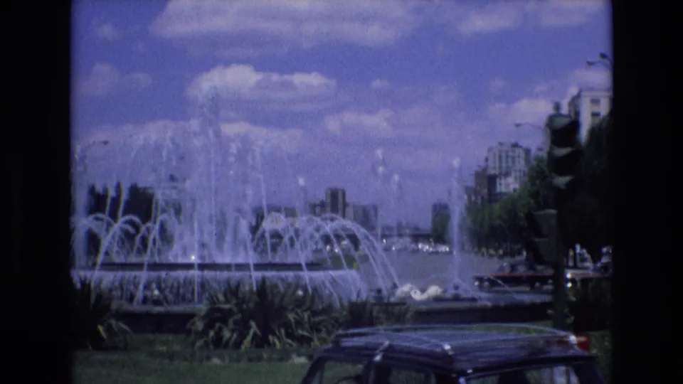 A 1972 Spain 8mm frame centers on the active fountain, with water jets rising above planted greenery and cars moving along the adjacent street.