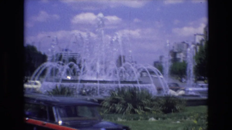 A 1972 Spain 8mm frame shows dense fountain spray in the foreground, a passing car at the lower edge, and urban buildings softened by home-movie focus.