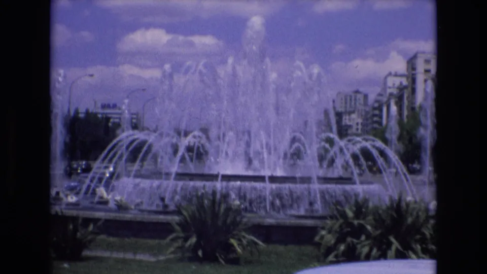 A 1972 Spain 8mm frame frames the fountain basin and tall central plume, with landscaped plants, roadside traffic, and apartment buildings around the urban park.