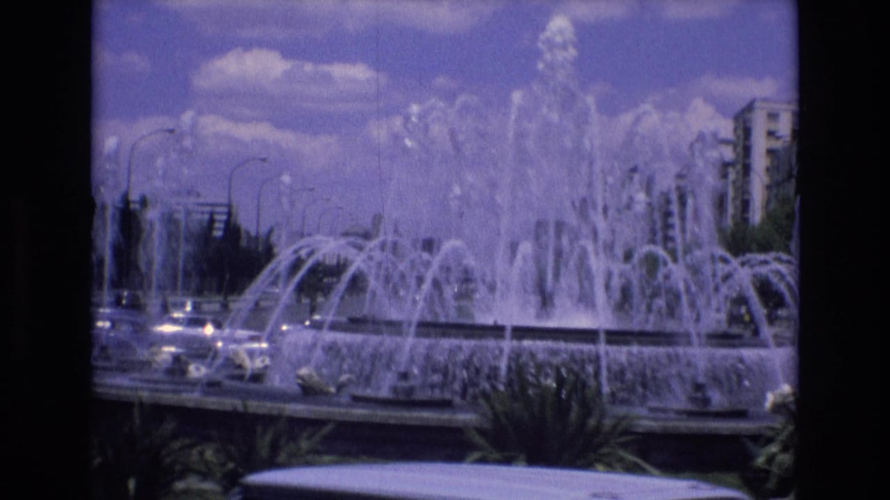 Spain 1972 home-movie frame showing a large public fountain with multiple water jets beside traffic, palm plantings, streetlights, and urban buildings.
