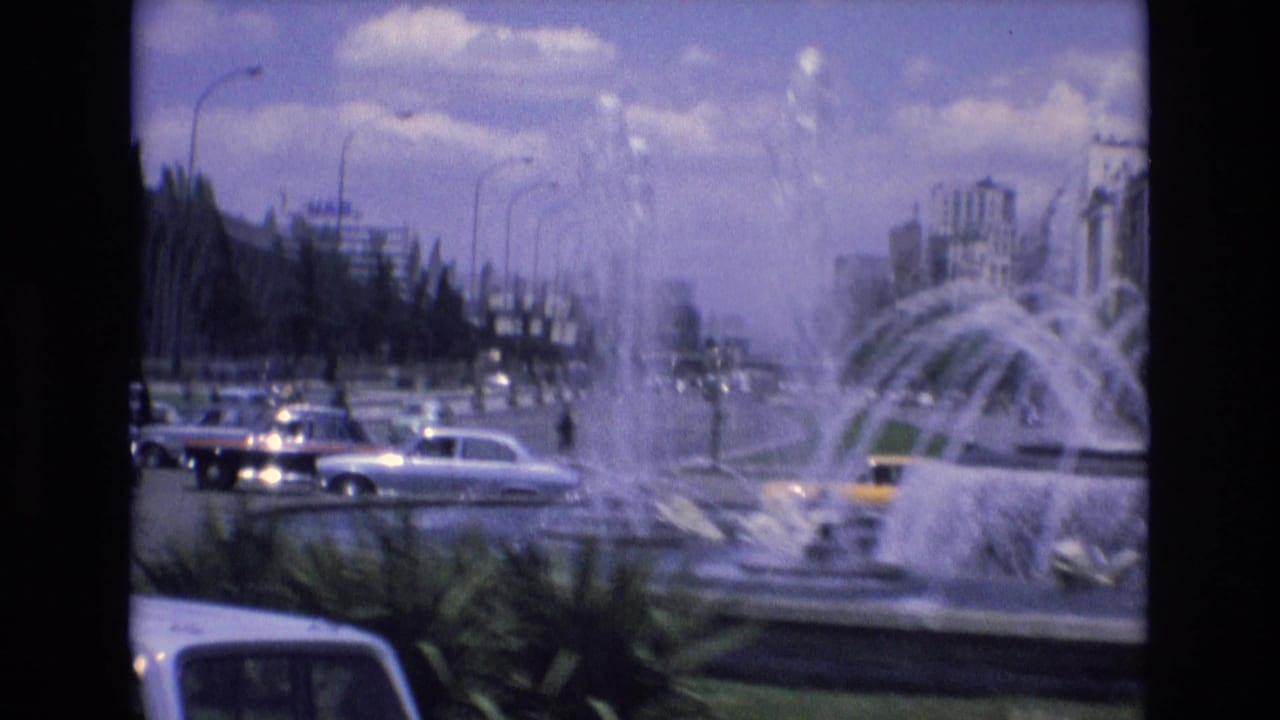 Cars pass a large fountain on a broad city road.