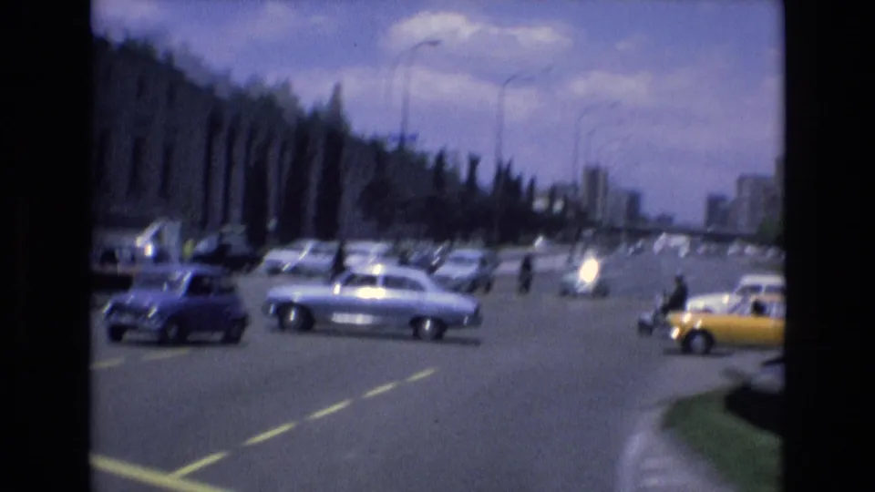 Spain 1972 home-movie frame showing a busy multi-lane city road with classic sedans, a yellow taxi, pedestrians at distance, and large urban buildings.