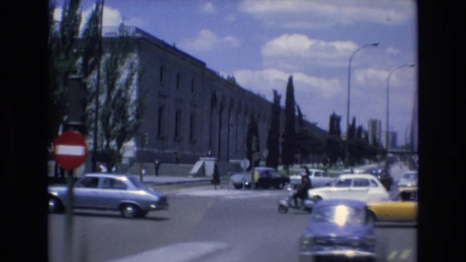Spain 1972 urban intersection with classic cars, a yellow taxi, a red no-entry sign, street trees, streetlights, and a long institutional-looking building.