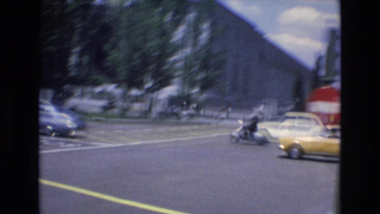 Wide city intersection with classic cars, a yellow taxi, trees, and large buildings.