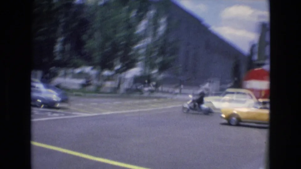 1972 Spain street view showing a broad intersection with classic cars, a yellow taxi, tree-lined sidewalks, and large urban buildings in soft 8mm focus.