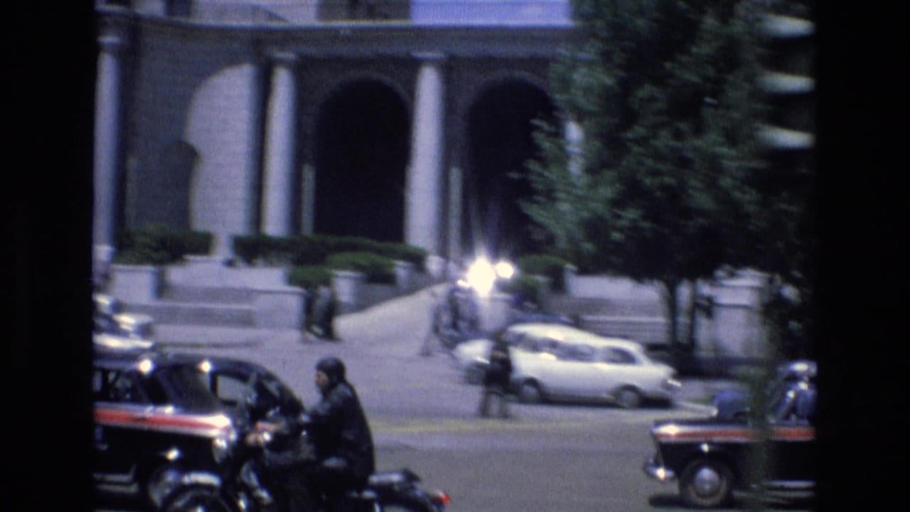 Motorcyclist rides through traffic near a columned building and passing cars.