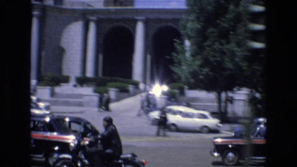 1972 Spain traffic scene with a motorcyclist moving past classic cars in front of a large building with columns and arched openings.