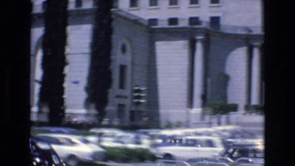 1972 Spain urban street frame showing rows of classic cars near a large pale building, tall trees, and columned architectural details.