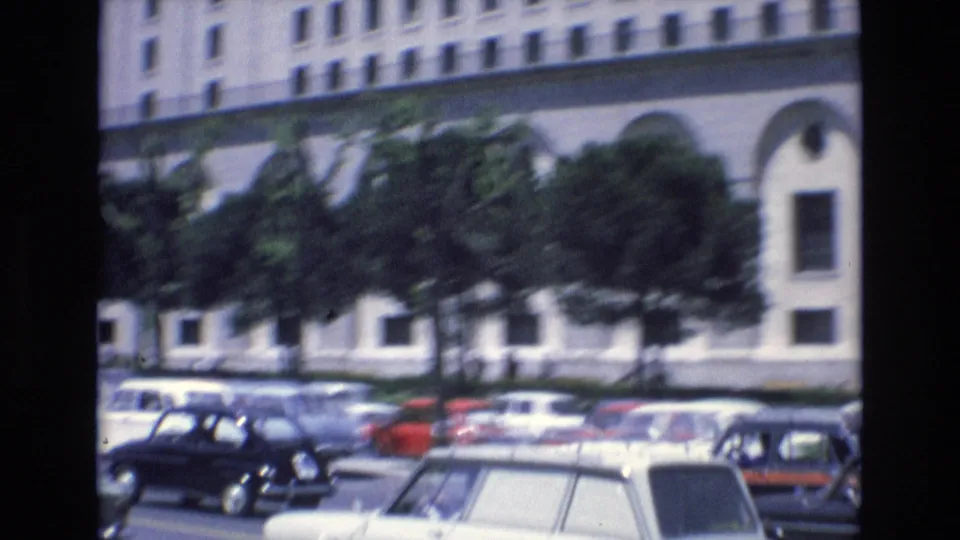 1972 Spain street traffic with compact classic cars, a light-colored station wagon, trees, and a large pale building in the background.