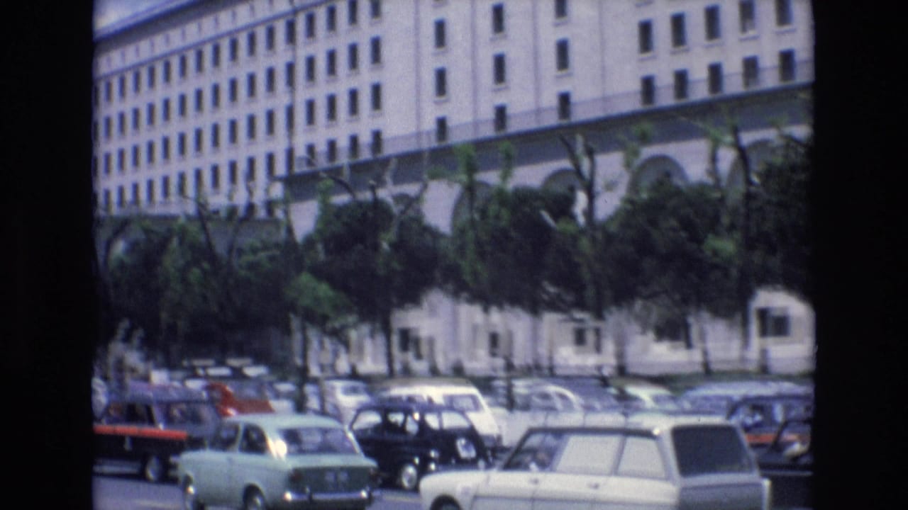 1972 Spain home-movie frame showing a broad urban street with classic cars, trees, and a large white building in the background.