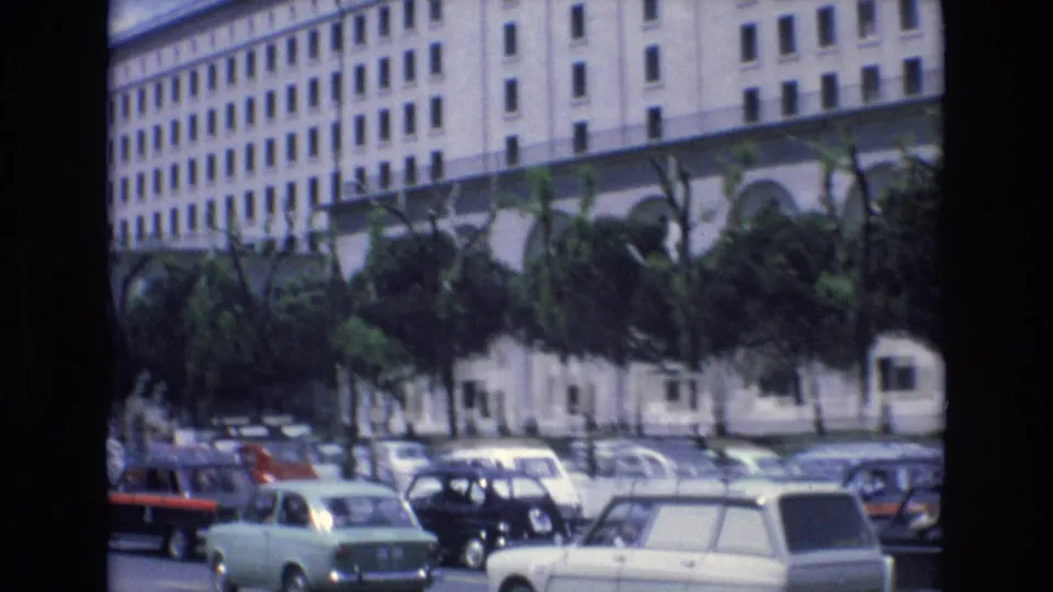 1972 Spain home-movie frame showing a broad urban street with classic cars, trees, and a large white building in the background.