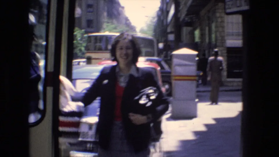 Adult woman in a dark jacket and red sweater stands near the open bus doorway on a sunlit Spanish city street in the 1972 home-movie footage.