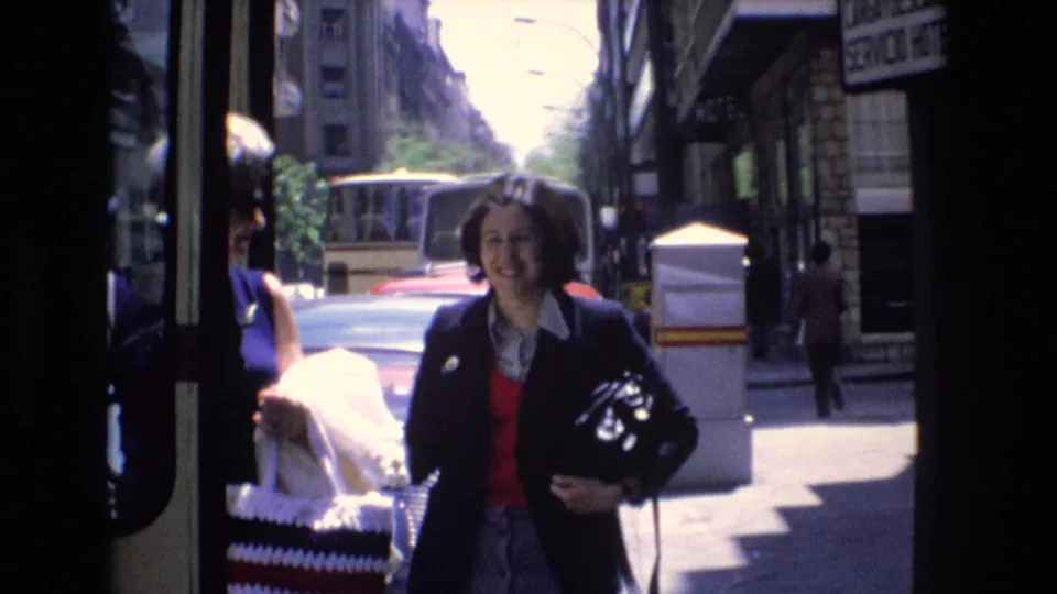 Adult woman carrying a dark handbag smiles toward the camera while standing beside the bus doorway on a sunny urban street in Spain, according to the supplied metadata.