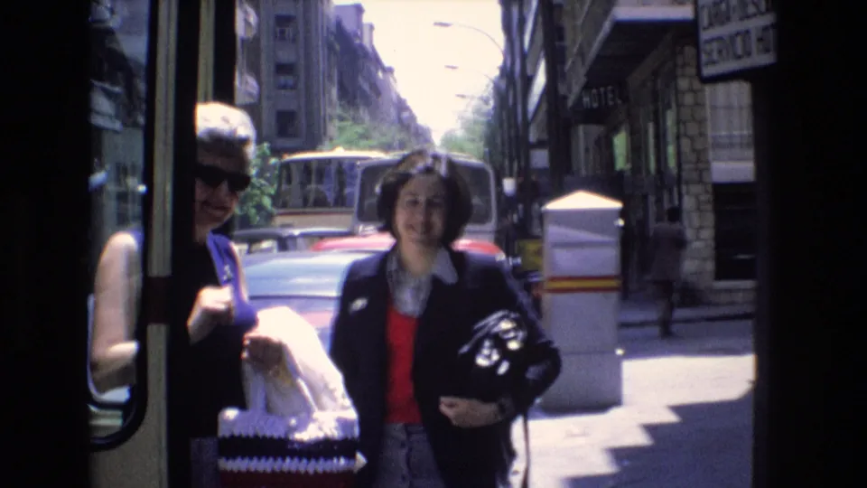 Two adult women are visible near the bus doorway, including one in sunglasses holding a patterned bag and another smiling toward the camera on a 1972 Spain city street.