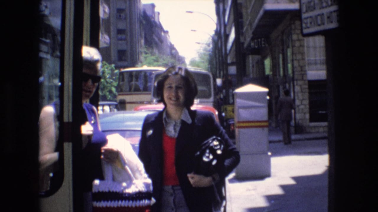 1972 Spain home-movie frame showing pedestrians near a shaded storefront, a woman carrying a helmet, and vehicles along a bright city street.