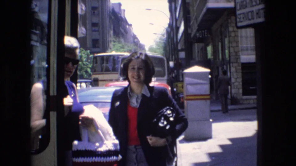 1972 Spain home-movie frame showing pedestrians near a shaded storefront, a woman carrying a helmet, and vehicles along a bright city street.