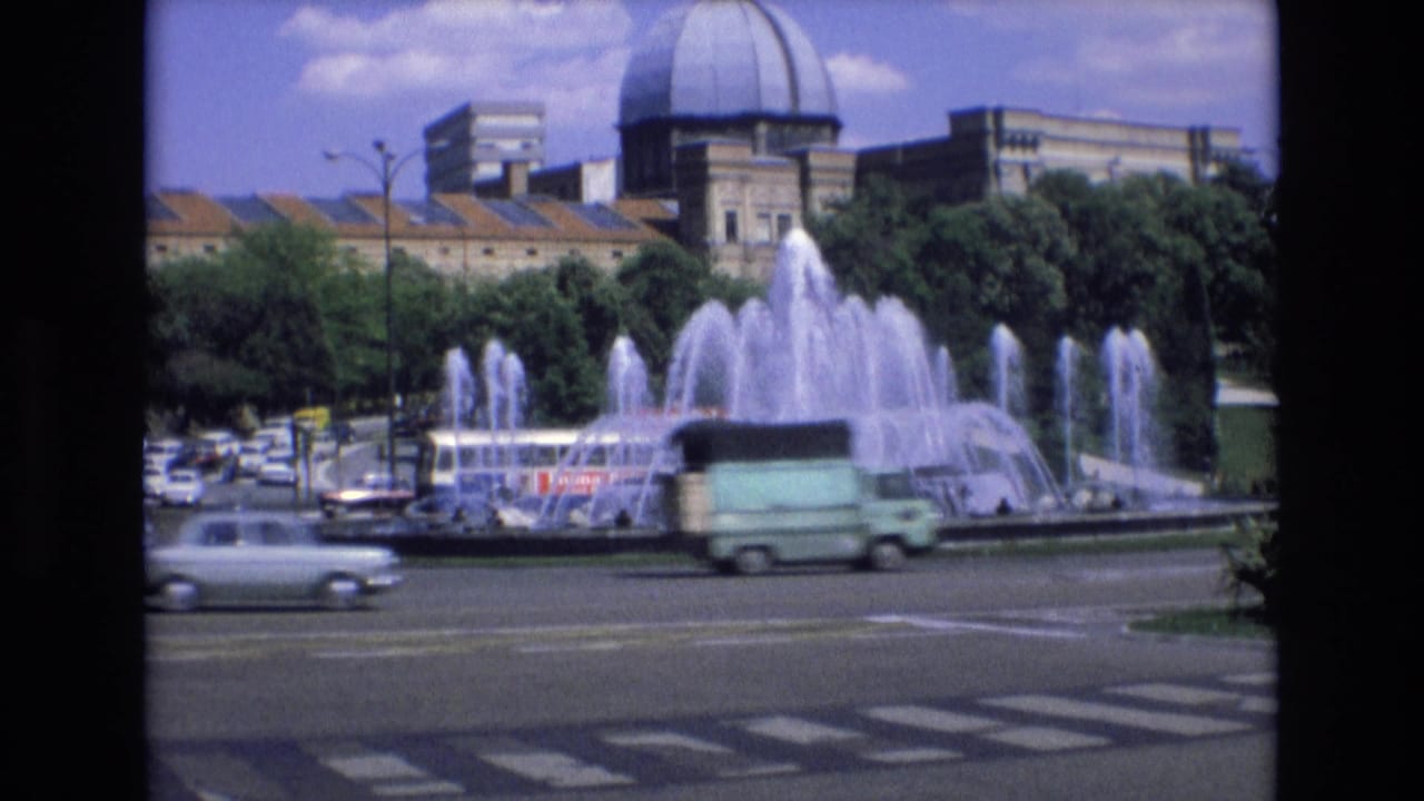 Large fountain beside a roadway with cars, trees, and a domed building in the background.