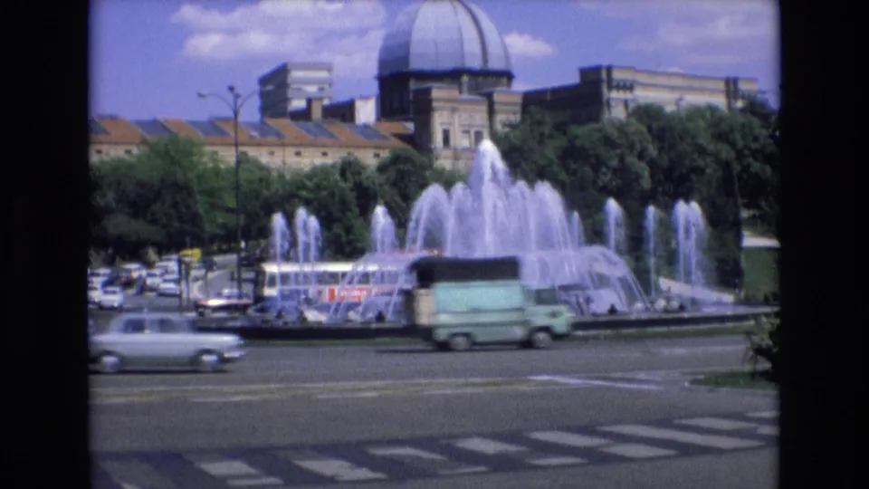 1972 Spain city-square view with a multi-jet fountain, moving cars, trees, and a prominent domed building behind the roundabout.