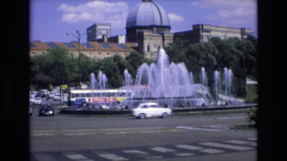 1972 Spain frame showing a public bus and several cars passing a large active fountain, with trees and a domed building behind it.