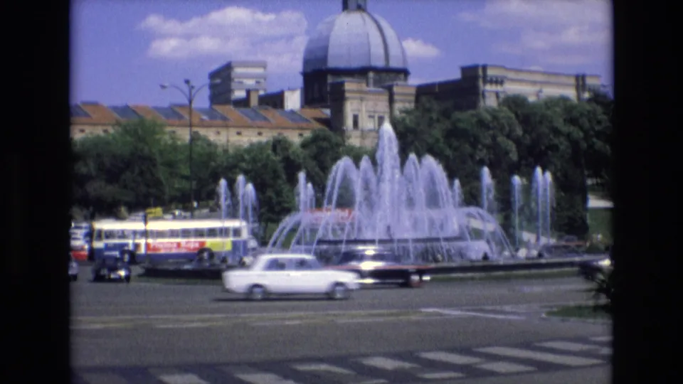 1972 Spain city-square frame with a white car, a public bus, active fountain jets, crosswalk markings, and a domed building in the background.