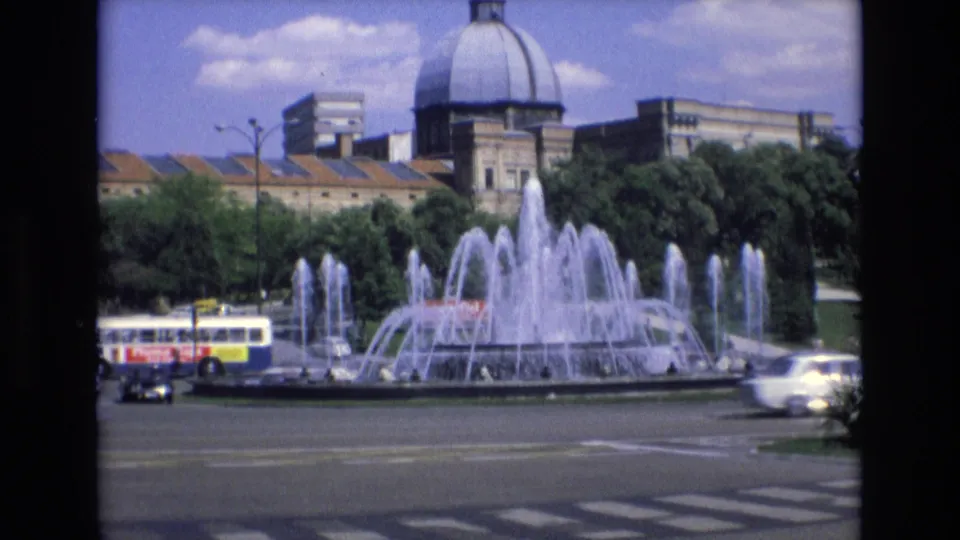 1972 Spain 8mm frame showing a large multi-jet fountain in a city roundabout, with cars, a bus, trees, and a domed building behind it.