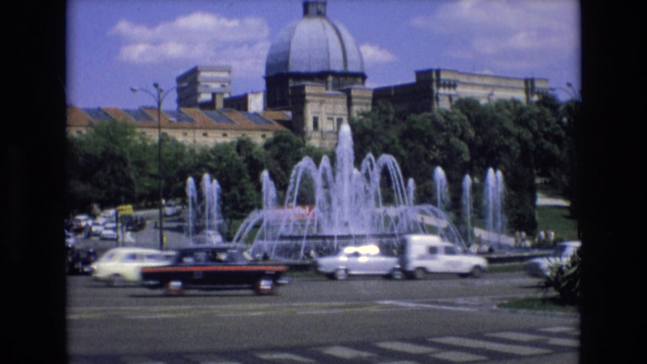Cars and a van pass in front of a large fountain and domed building.