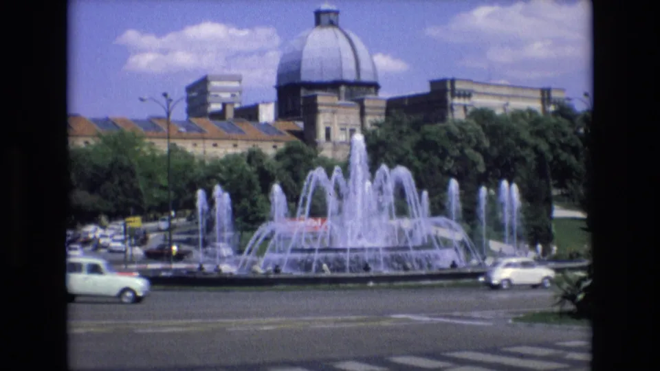1972 Spain 8mm frame looking across a road and crosswalk toward a large public fountain with trees and a domed building in the background.