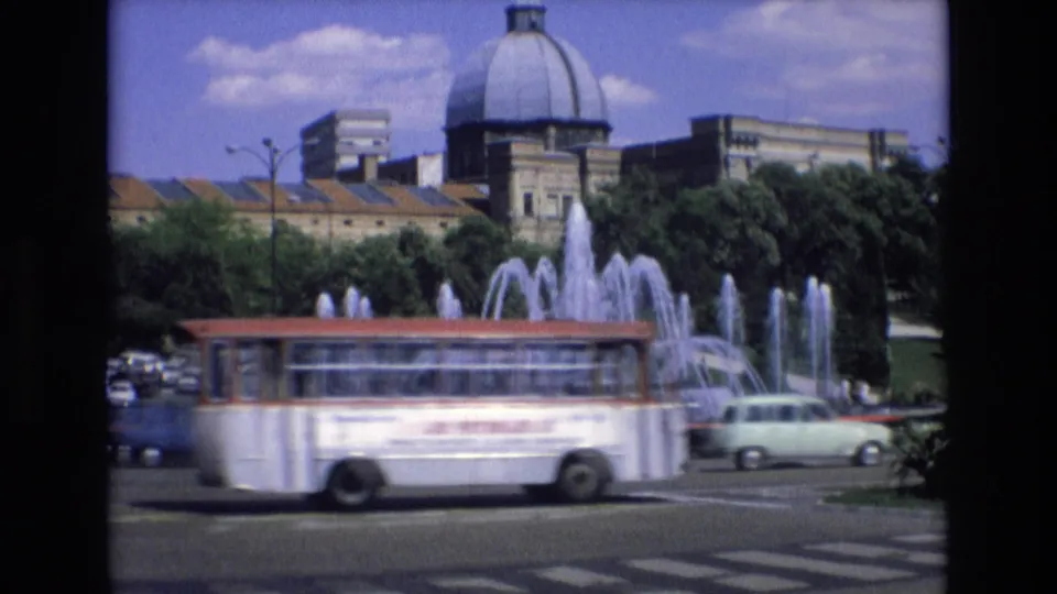 1972 Spain 8mm frame showing a white public bus with red trim moving across the foreground, partially obscuring the large fountain and domed building behind it.