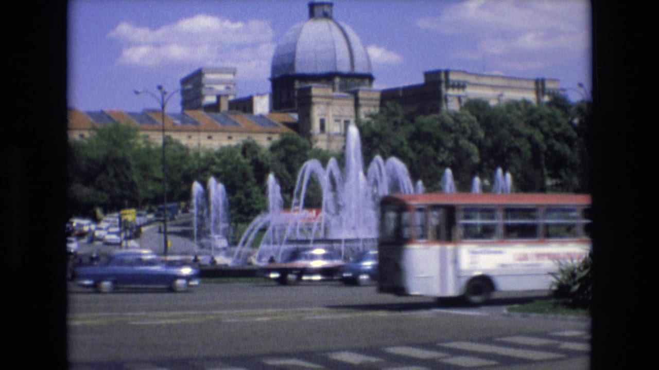 Spain 1972 8mm frame showing a sunlit city square with arcing fountain jets, classic cars, a passing bus, trees, and a prominent domed building beyond the roadway.