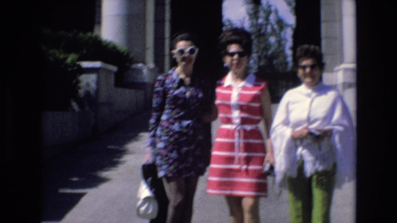 Three women wearing sunglasses walk toward the camera beside columns and a shaded building entrance.