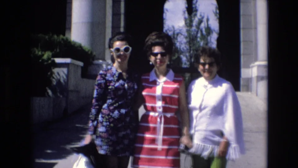 Spain 1972 8mm frame showing three women smiling and posing for the camera in bright 1970s clothing beside columns and a shaded walkway in an urban setting.
