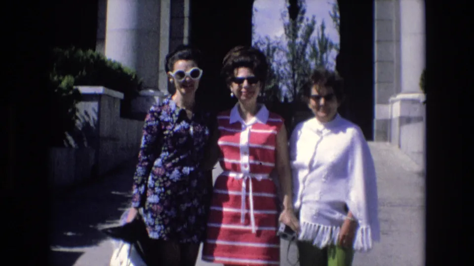 Three adult women in Spain in 1972 continue posing on a sunlit walkway, with the left woman leaning slightly and the columned building facade behind them.
