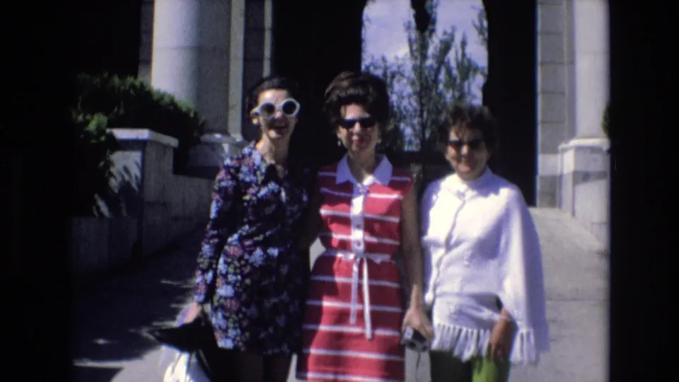 Three adult women in 1972 Spain remain posed close together before a columned building, showing clear faces, sunglasses, handbags, and 1970s-style clothing.