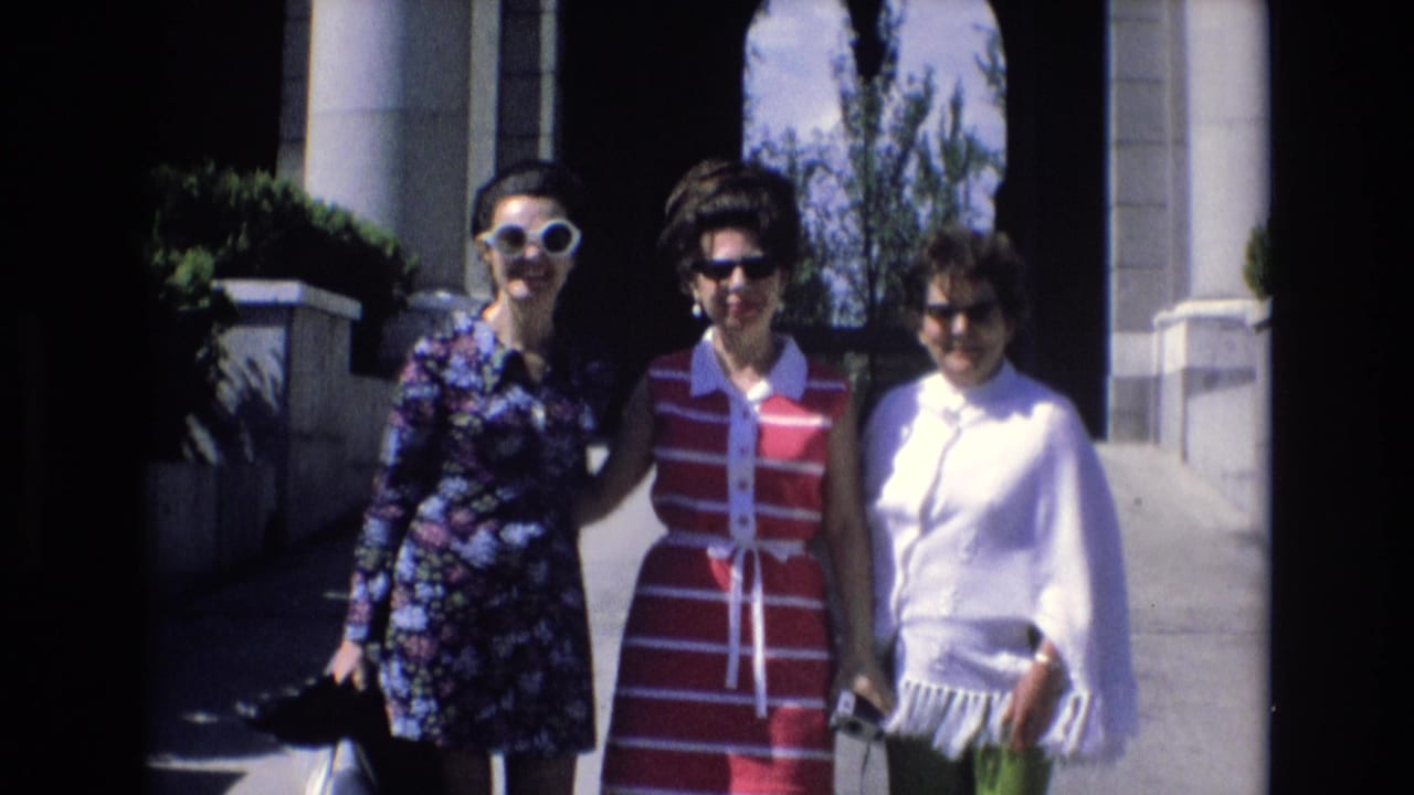 Three women in 1970s clothing pose outdoors in Spain beside columns and an arched building entrance, with trees and bright sky behind them.
