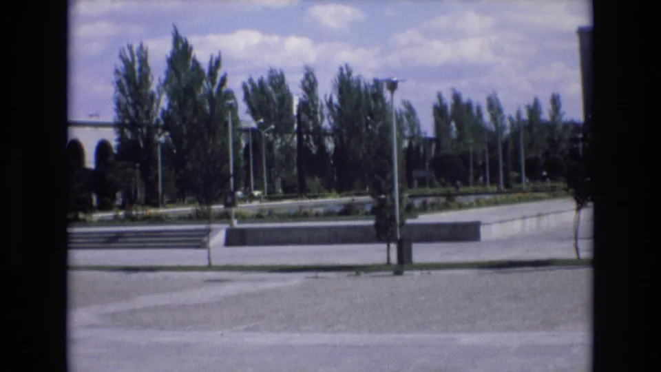 A wide 8mm view in Spain shows trees, lampposts, concrete walkways, low steps, and part of a colonnaded building under a bright sky.