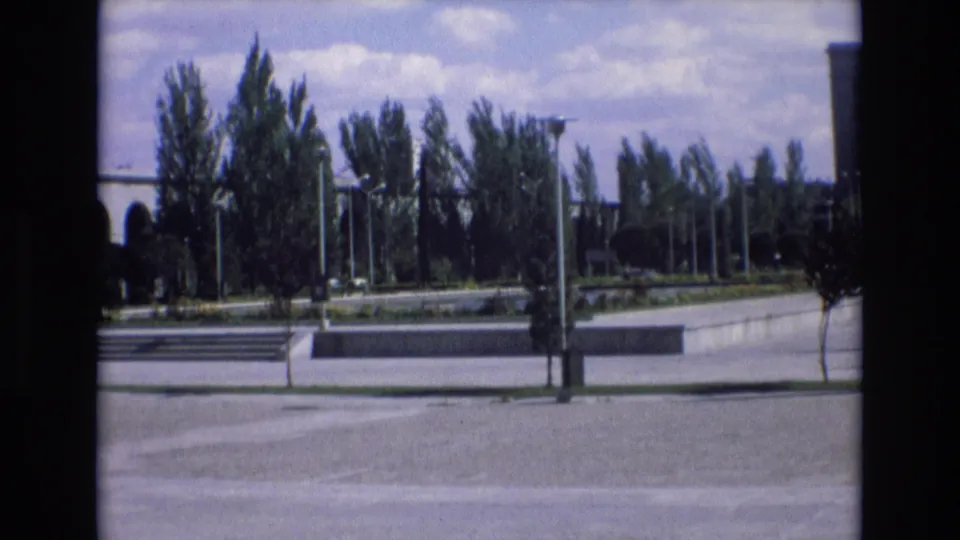 A handheld wide shot shows a public landscaped area in Spain with windblown trees, lampposts, concrete steps, and open paving.