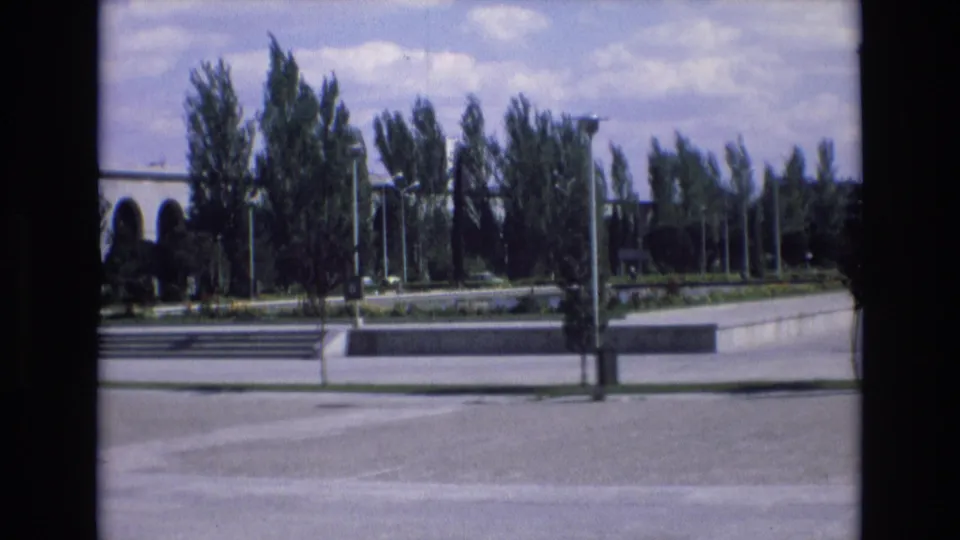 The final frame holds on the same public exterior in Spain, with trees, lampposts, low concrete steps, and a paved foreground captured on 8mm film.