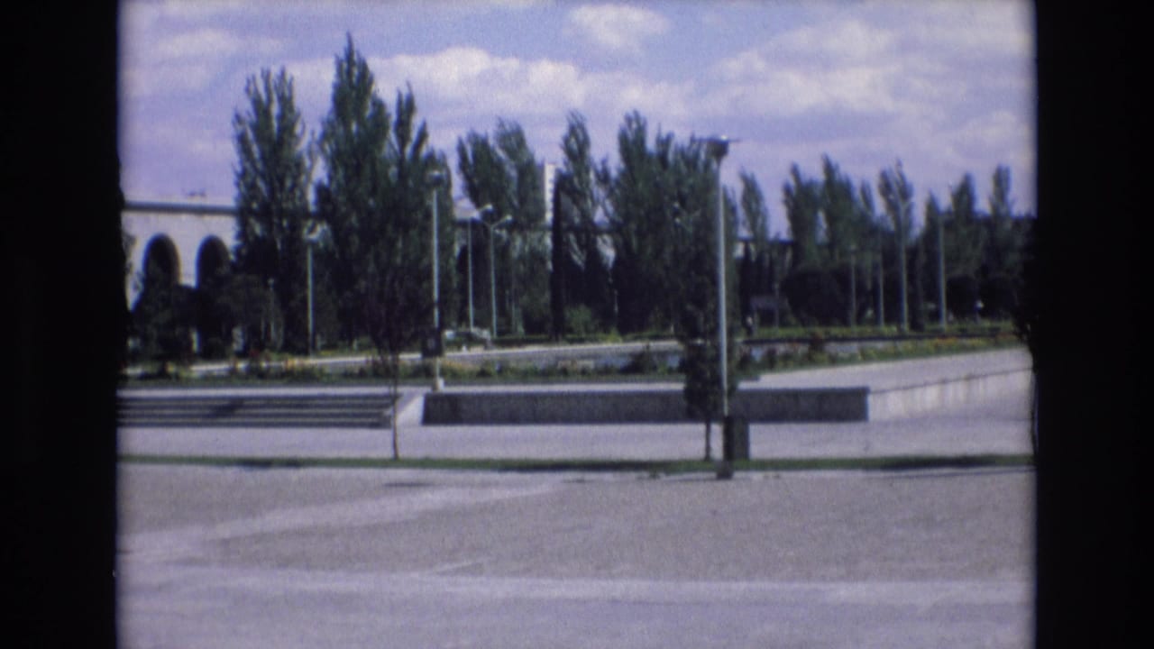 1972 Spain home-movie frame showing a broad paved plaza with tall trees, modernist arches at left, and a bright partly cloudy sky.